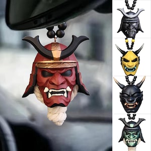 May include: A red and black samurai mask car charm with a white rope and black beads. The mask has a fierce expression and is part of a collection of similar masks in black, yellow, and green.