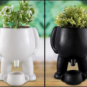 Pode incluir: Duas floreiras extravagantes, uma branca e outra preta, cada uma em forma de figura estilizada com um pequeno vaso por baixo. A floreira branca contém uma planta verde e branca, a preta uma planta verde. Um artigo decorativo único.