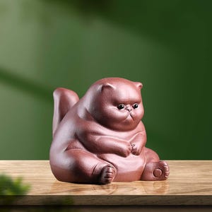 Può includere: Statuetta di gatto in ceramica marrone, seduta. Il gatto ha un corpo rotondo, piccole orecchie e un'espressione accigliata. La statuetta è su una superficie di legno con sfondo verde.