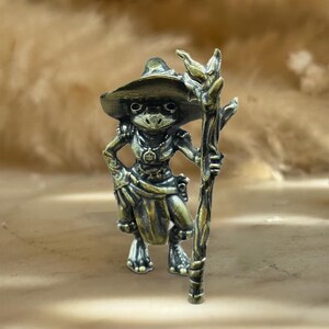 May include: A bronze frog figurine, wearing a hat and tunic, holding a staff. The frog has a detailed face and is posed with one hand on its hip. The staff has a detailed, textured design. The figurine is a fantasy-themed item.