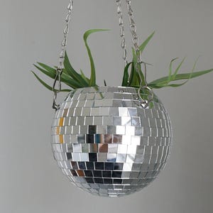 May include: A silver disco ball planter, suspended by a chain, reflects light. The spherical planter is covered in small, mirrored squares. Green plants sprout from the top, adding a touch of nature to the decorative piece.
