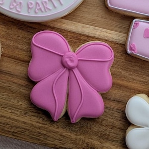 May include: A close-up of decorated cookies on a wooden surface. The central cookie is shaped like a pink bow with detailed icing. Other cookies include a flower and a purse shape, all with pink and white icing.