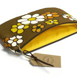 May include: A brown zippered pouch with a yellow lining. The pouch is decorated with a floral pattern of white, yellow, and orange flowers. The pouch has a brown leather tag with the text "Ella&Olive" printed on it.