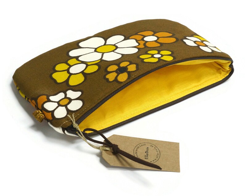 May include: A brown zippered pouch with a yellow lining. The pouch is decorated with a floral pattern of white, yellow, and orange flowers. The pouch has a brown leather tag with the text "Ella&Olive" printed on it.