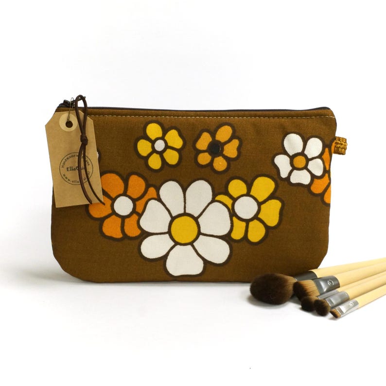 May include: Brown fabric makeup bag with a floral pattern of white, yellow, and orange daisies. The bag has a zipper closure and a brown leather tag with the text "Ella Grace" on it.