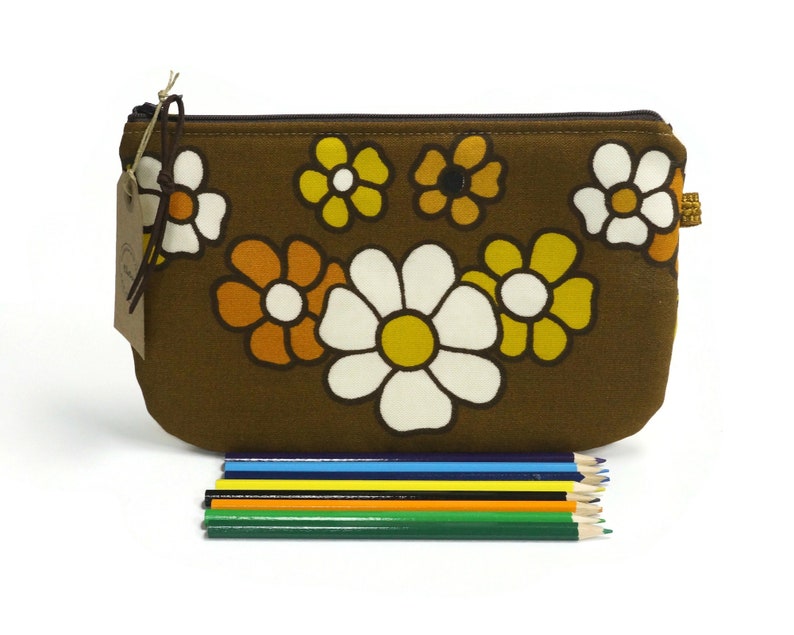 May include: A brown fabric zippered pouch with a floral pattern featuring white, yellow, and orange daisies. The pouch is lying on a white surface with a set of colored pencils in front of it.