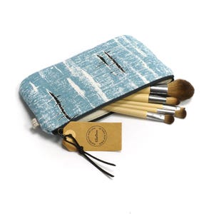 Può includere: Una trousse per il trucco con motivo blu e bianco con chiusura a zip. La trousse è aperta e contiene un set di pennelli per il trucco con manici in legno.