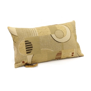 Peut inclure: Un coussin rectangulaire en tissu texturé dans des tons beige, ocre et marron. La surface présente un motif géométrique composé de cercles, de demi-cercles et de blocs rayés. Une petite étiquette en tissu beige est fixée au coin inférieur.