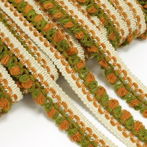 Könnte beinhalten: Nahaufnahme einer dekorativen Borte mit sich wiederholendem Muster. Die Borte weist abwechselnd cremefarbene, grüne und orangefarbene Streifen auf. Die grünen und orangenen Abschnitte haben ein strukturiertes, geschlungenes Design. Diese Borte eignet sich für verschiedene Bastel- und Nähprojekte.