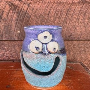 Puede incluir: Jarrón de cerámica hecho a mano con un diseño de cara caprichoso. El jarrón presenta una combinación de colores azul y morado, con tres detalles en forma de ojo y una boca grande sonriente. La superficie texturizada se suma a su atractivo artístico único.