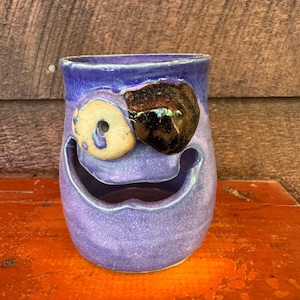 May include: A handmade ceramic mug in shades of purple, featuring a whimsical face design. The mug has a large, smiling mouth and two eye-shaped embellishments, one beige and one dark brown. The mug is sitting on an orange surface.