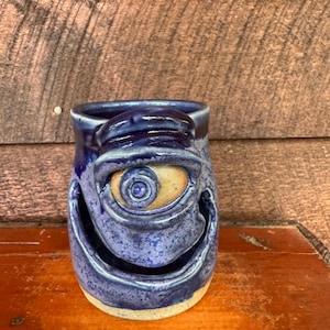 May include: A ceramic mug with a glossy blue glaze featuring a sculpted, singular large eye in the center and a wide, open mouth carved into the front. The base and the area around the eye reveal a natural, light tan clay color beneath the glaze.