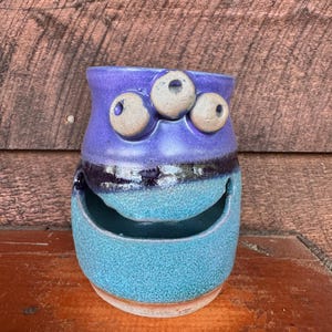 May include: A handmade ceramic vase with a whimsical face design. The vase features a purple top with three beige, eye-shaped protrusions, a black band, and a turquoise body with a smiling mouth-shaped opening. The base is unglazed.