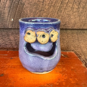 May include: A handmade ceramic mug in shades of purple, featuring a whimsical face design. The mug has three circular eye-like protrusions and a wide, smiling mouth. The glaze gives a textured appearance, adding to its unique, artistic appeal.