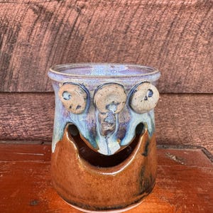May include: A handmade ceramic vessel with a smiling face design. The top rim is a light blue with three circular accents. The body transitions from blue to brown, with a large, curved opening forming the mouth. The piece is set against a wooden background.