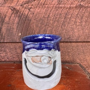 May include: A handmade ceramic mug with a whimsical face design. The mug features a blue rim, a white body, and a smiling face with an eye and mouth carved into the surface. The mug is approximately 4 inches tall.