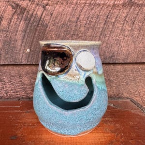 May include: A whimsical, handmade ceramic vase with a face design. The vase features a turquoise body, a brown and white eye, and a smiling mouth opening. The top rim is a light tan color. The vase is sitting on a wooden surface.
