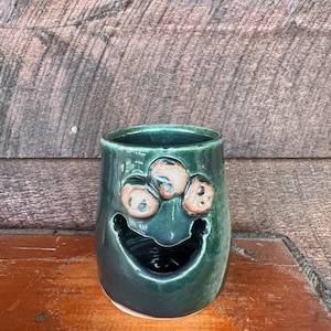 Puede incluir: Una taza de cerámica verde hecha a mano con un diseño de cara caprichoso. La taza presenta tres acentos circulares de color canela sobre una boca ancha y sonriente. El interior de la taza es oscuro y el exterior tiene un acabado brillante.