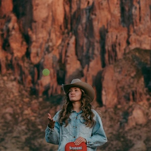 Puede incluir: Una mujer con una chaqueta vaquera y un sombrero de vaquero sostiene una raqueta de paddleball roja con un dise&ntilde;o amarillo y negro. La raqueta tiene la palabra "Neonami" impresa en ella. La mujer est&aacute; de pie frente a una formaci&oacute;n rocosa roja.