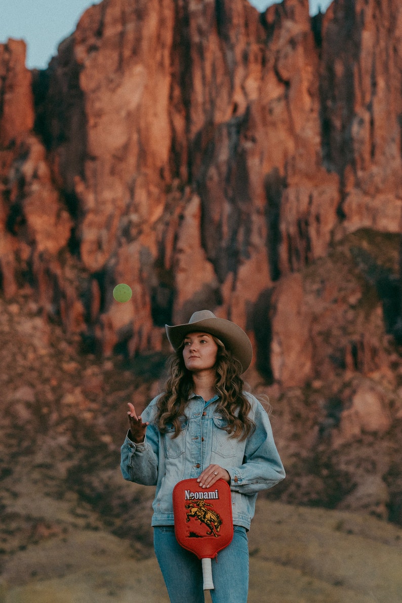 Puede incluir: Una mujer con una chaqueta vaquera y un sombrero de vaquero sostiene una raqueta de paddleball roja con un dise&ntilde;o amarillo y negro. La raqueta tiene la palabra "Neonami" impresa en ella. La mujer est&aacute; de pie frente a una formaci&oacute;n rocosa roja.
