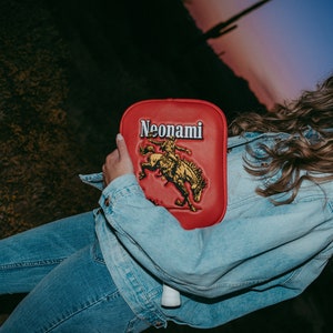 Puede incluir: Una mujer con una chaqueta vaquera y un sombrero de vaquero sostiene una paleta roja con la palabra "Neonami" y un gr&aacute;fico de un vaquero montando un caballo.
