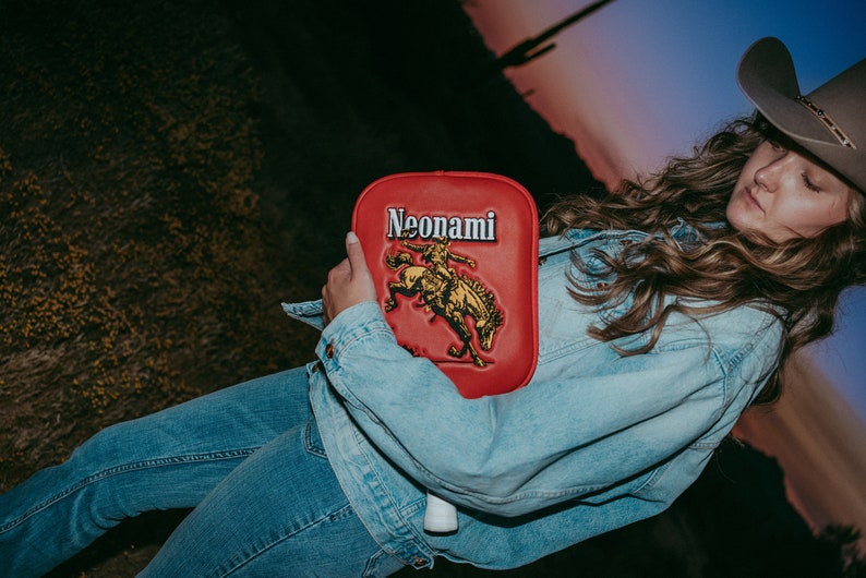 Puede incluir: Una mujer con una chaqueta vaquera y un sombrero de vaquero sostiene una paleta roja con la palabra "Neonami" y un gr&aacute;fico de un vaquero montando un caballo.
