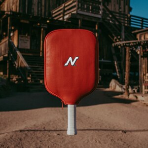 Puede incluir: Una pala de pickleball roja con un logotipo "N" blanco y un mango blanco.