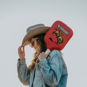 Puede incluir: Una mujer con una chaqueta vaquera y un sombrero de vaquero marr&oacute;n sostiene una paleta roja con una imagen amarilla de un vaquero montando un caballo. La paleta tiene el texto "Neonami" impreso en ella.