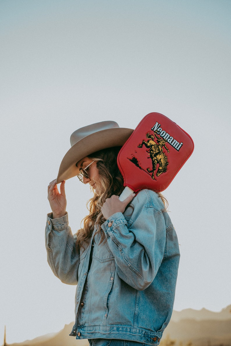 Puede incluir: Una mujer con una chaqueta vaquera y un sombrero de vaquero marr&oacute;n sostiene una paleta roja con una imagen amarilla de un vaquero montando un caballo. La paleta tiene el texto "Neonami" impreso en ella.