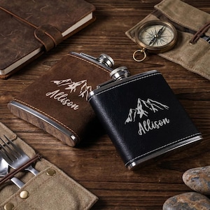 Könnte beinhalten: Zwei personalisierte Flachmänner, einer braun und einer schwarz, mit einem Bergdesign und dem eingravierten Namen "Allison". Die Flachmänner haben einen Edelstahlrand und -verschluss. Ein Kompass, ein Notizbuch und ein Besteckset sind ebenfalls zu sehen.