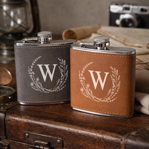 Puede incluir: Dos petacas de cuero, una gris y otra marrón, con tapas plateadas. Cada petaca presenta un diseño de corona floral blanca alrededor de una gran letra blanca "W". Las petacas están sobre una superficie de madera.