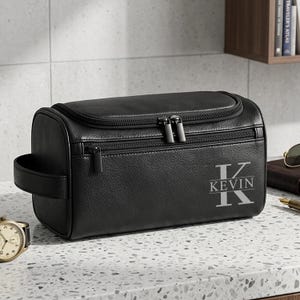 May include: Black leather wash bag with a top zip and a side handle. The bag has the name "KEVIN" in a stylized font. A watch and sunglasses are visible in the background.