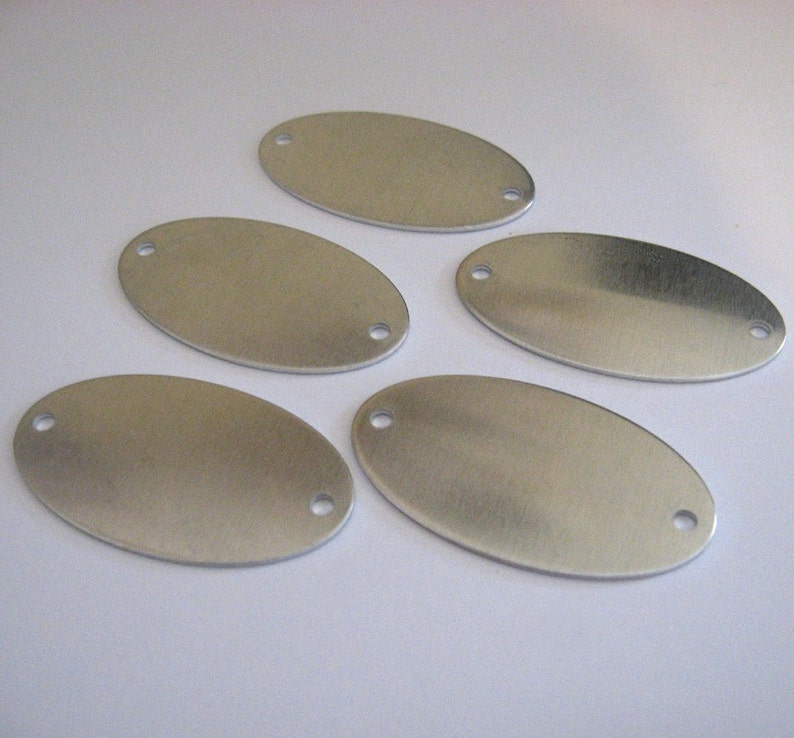 May include: Five silver oval-shaped metal tags with two holes each. The tags are blank and ready for engraving.