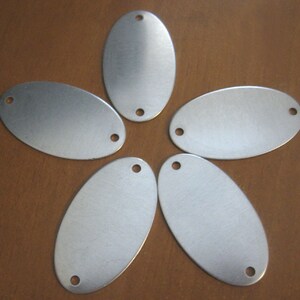 May include: Five silver oval-shaped metal tags with two holes each. The tags are arranged in a star shape on a brown wooden surface.