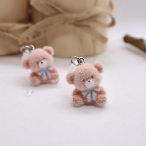 Könnte beinhalten: Zwei kleine, rosa Teddybär-Ohrringe mit silberfarbenen Haken. Jeder Bär hat eine hellblaue Schleife und eine flauschige Textur. Die Ohrringe sind vor einem weichen, weißen Hintergrund.