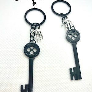 Könnte beinhalten: Zwei schwarze Schlüsselanhänger mit einem Skelett-Hand-Anhänger. Die Schlüssel haben eine kreisförmige Oberseite mit drei Löchern und einen schlüsselförmigen Boden. Das Wort "Coraline" ist oben in Schreibschrift geschrieben.