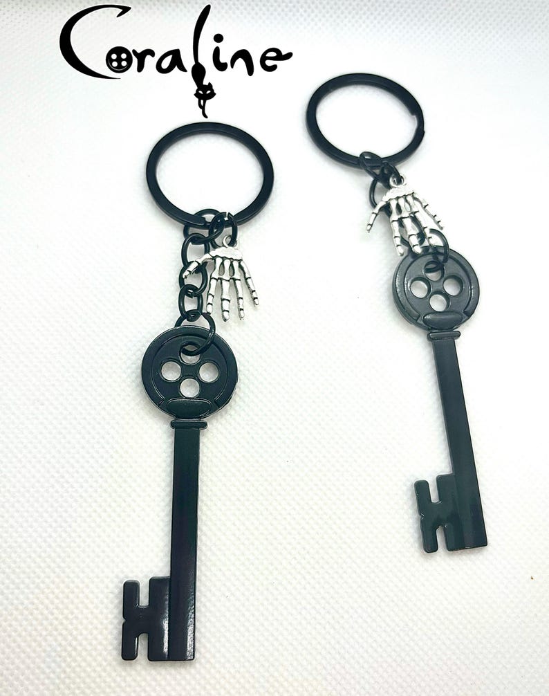 Op de afbeelding: Twee zwarte sleutelhangers met een skelet hand bedel. De sleutels hebben een cirkelvormige bovenkant met drie gaten en een sleutelvormige onderkant. Het woord "Coraline" staat in cursief bovenaan.