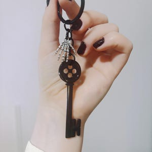 Op de afbeelding: Zwarte sleutelhanger in de vorm van een sleutel met een skelet hand bedel. De sleutel heeft een cirkelvormige bovenkant met vier gaten en een lange, rechthoekige steel. De sleutelhanger is zwart.
