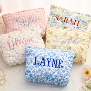 May include: Five quilted cosmetic bags in various floral and gingham patterns. Each bag is embroidered with a different name in a contrasting color: Kaylee, Olivia, Layne, Sarah, and Layla. The bags are in shades of pink, blue, and yellow.