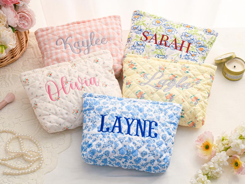 May include: Five quilted cosmetic bags in various floral and gingham patterns. Each bag is embroidered with a different name in a contrasting color: Kaylee, Olivia, Layne, Sarah, and Layla. The bags are in shades of pink, blue, and yellow.