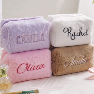 May include: Four fluffy cosmetic bags in pastel shades: lavender, white, pink, and tan. Each bag is embroidered with a name in a matching colour. The rectangular bags have a zip closure, ideal for storing makeup and beauty products.