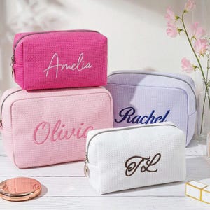 May include: Four rectangular cosmetic bags with a textured waffle-weave fabric and silver zips. The bags are in hot pink with the name Amelia, light pink with Olivia, lavender with Rachel, and white with the embroidered initials TL.