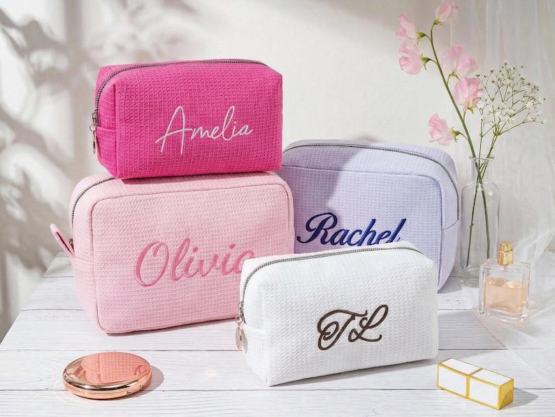 May include: Four rectangular cosmetic bags with a textured waffle-weave fabric and silver zips. The bags are in hot pink with the name Amelia, light pink with Olivia, lavender with Rachel, and white with the embroidered initials TL.