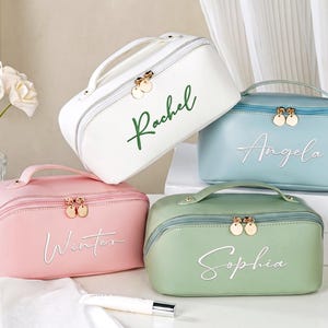 May include: Four rectangular cosmetic bags with top handles and gold-toned double zipper pulls. The white bag features the name Rachel in green, the blue bag says Angela, the pink bag says Winter, and the green bag says Sophia in white script.