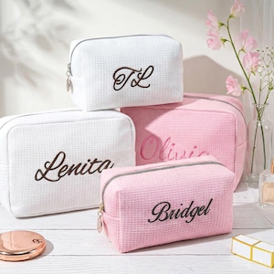 May include: Three rectangular cosmetic bags with a waffle-textured fabric. Two bags are white with dark brown embroidered names Lenita and TL. Two bags are light pink with dark brown embroidered names Olivia and Bridgel. Each bag has a silver zipper.