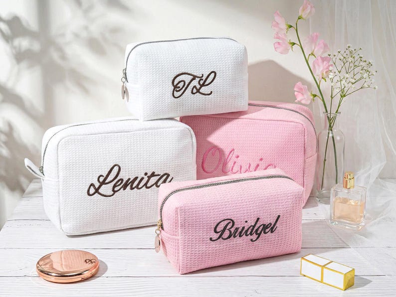 May include: Three rectangular cosmetic bags with a waffle-textured fabric. Two bags are white with dark brown embroidered names Lenita and TL. Two bags are light pink with dark brown embroidered names Olivia and Bridgel. Each bag has a silver zipper.