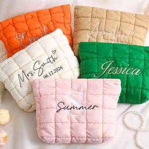 Op de afbeelding: Een verzameling gewatteerde make-uptassen in verschillende kleuren: oranje, beige, groen, wit en roze. Elke tas is geborduurd met namen of initialen in een sierlijk lettertype. De tassen zijn gerangschikt op een wit oppervlak, wat duidt op een cadeau of gepersonaliseerd item.