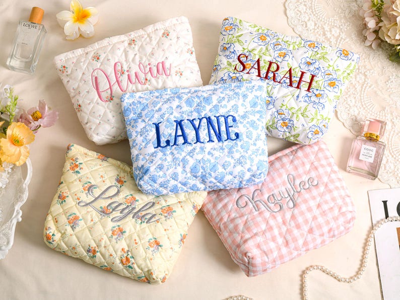 May include: Five quilted cosmetic bags with embroidered names. The bags feature floral and gingham patterns in white, blue, yellow, and pink. Names embroidered include "Olivia", "Sarah", "Layne", "Layla", and "Kaylee".