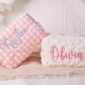 May include: Two quilted cosmetic bags. One is pink and white gingham with "Kaylee" embroidered in light blue. The other is off-white with a floral pattern and "Olivia" embroidered in pink. Both have a zipper closure.
