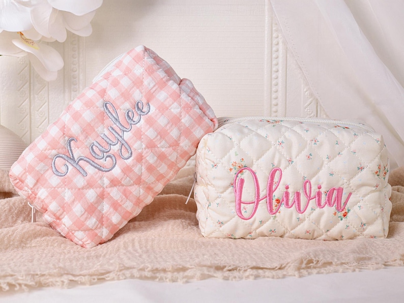 May include: Two quilted cosmetic bags. One is pink and white gingham with "Kaylee" embroidered in light blue. The other is off-white with a floral pattern and "Olivia" embroidered in pink. Both have a zipper closure.